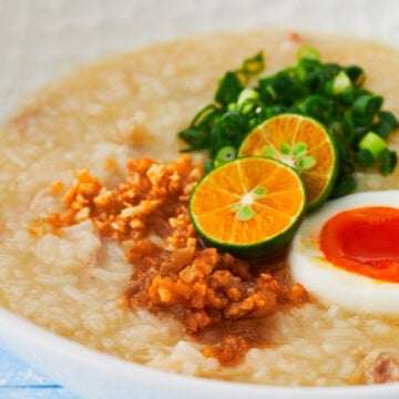 This mouthwatering bowl of Arroz Caldo (Filipino Chicken and Rice Porridge) is garnished with chicken cracklins, crispy garlic, a soft boiled egg, scallions, and Calamansi.