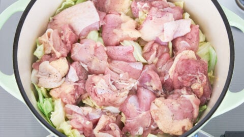 Salt and pepper seasoned chicken thighs on a bed of cabbage in a green dutch oven for making chicken stew.