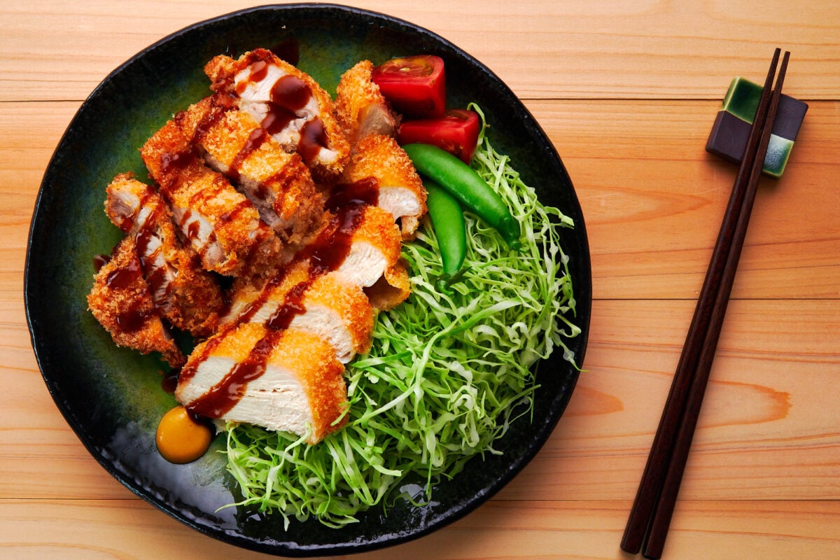 Crispy Chicken Katsu drizzled with katsu sauce on a green plate with shredded cabbage, tomatoes, and snap peas.