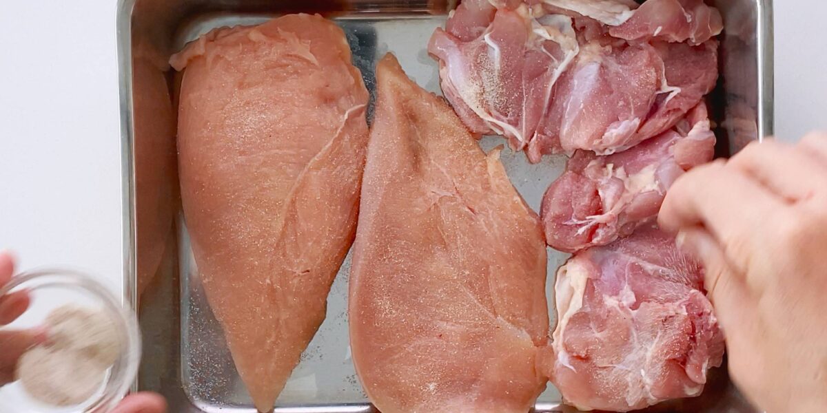 Seasoning chicken in a metal tray with salt and pepper.