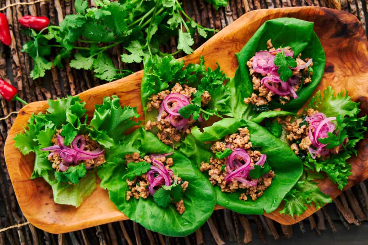 Overhead shot of Asian Chicken Lettuce Wraps beautifully arranged on a natural wood platter, surrounded by fresh chili peppers and cilantro.