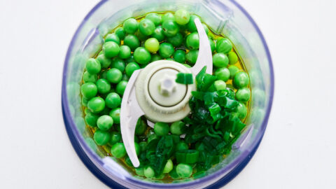 Blanched green peas, scallions, and olive oil in a blender on a white counter. For making Green Pea Pesto.