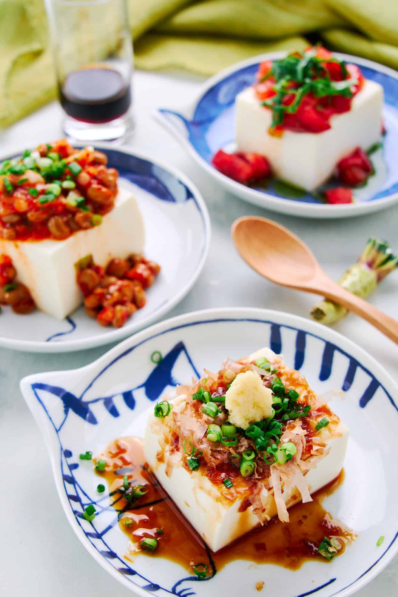 Hiyayakko is a Japanese cold tofu dish topped with katsuobushi, scallions, ginger, and soy sauce. In the background, two modern variations of this tofu salad include Kimchi Natto and Caprese Salad.