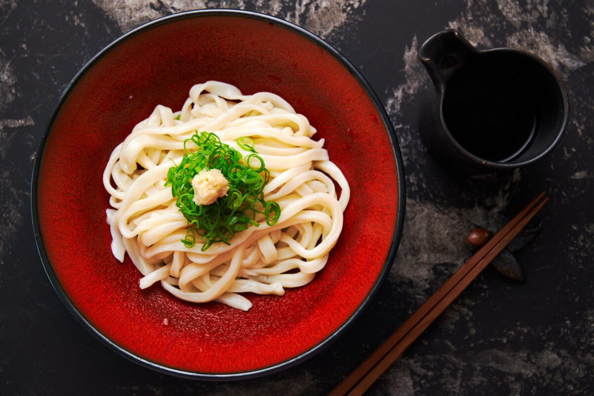 Red plate with a nest of chilled udon noodles garnished with scallions and ginger.