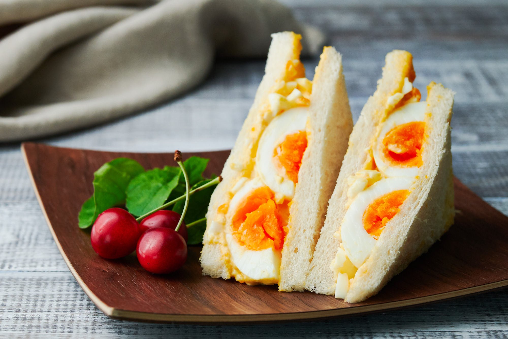 Japanese style egg sandwich including whole eggs on a wooden plate with cherries and arugula.