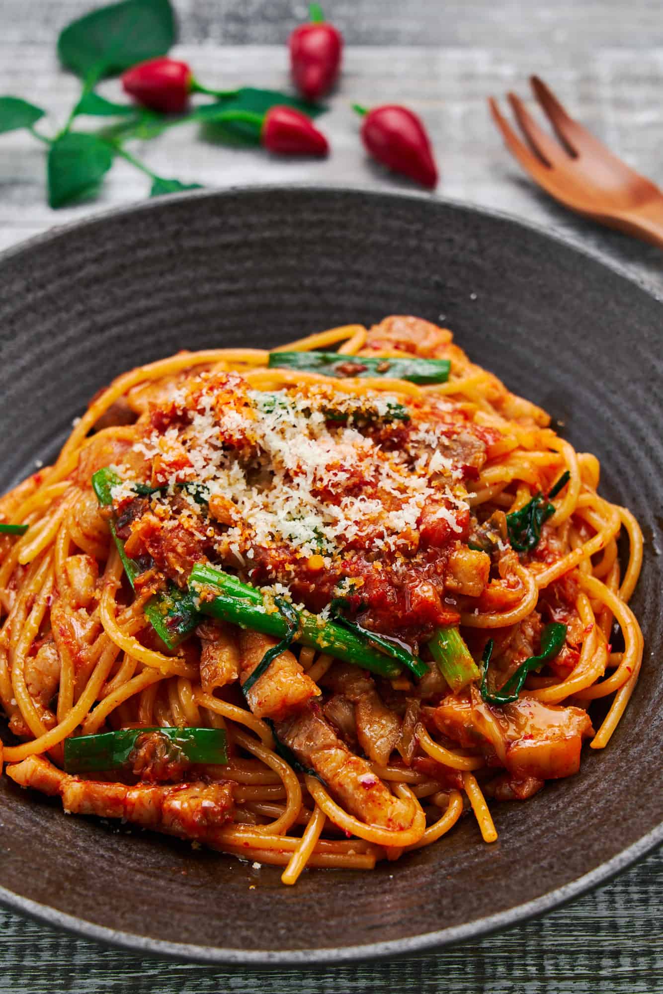 Spicy and savory 15 minute Pork & Kimchi Pasta piled high on a dark plate, the vibrant red of the kimchi and juicy pork belly pops against the creamy strands of spaghetti and vibrant green scallions.