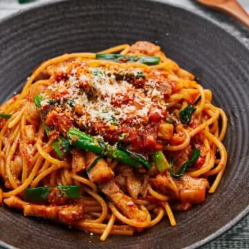 Spicy and savory 15 minute Pork & Kimchi Pasta piled high on a dark plate, the vibrant red of the kimchi and juicy pork belly pops against the creamy strands of spaghetti and vibrant green scallions.