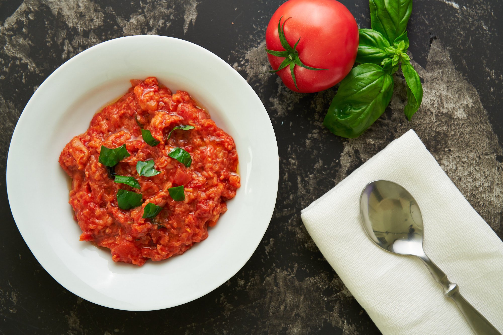 Pappa al Pomodoro (Tuscan Bread and Tomato Soup) in a white bowl on a dark surface with a fresh tomato and basil leaves.