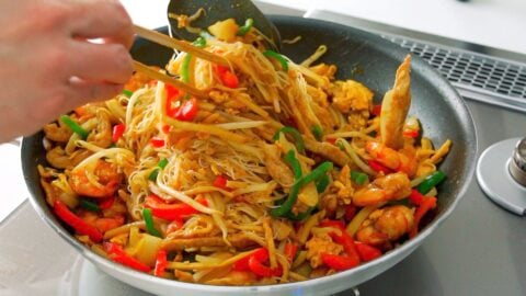 Tossing Singapore Noodles in a frying pan with chopsticks and a spatula.