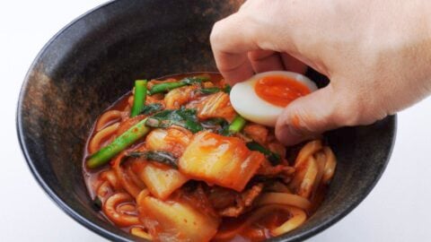 Setting a soft boiled egg on top of a bowl of spicy kimchi udon noodles.