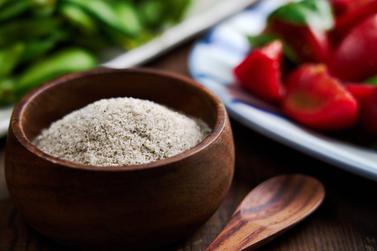 Wooden bowl filled with umami salt with edamame and tomatoes in the background.