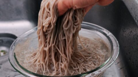 Rinsing boiled soba noodles under running water to remove excess starch from the surface of the noodles.